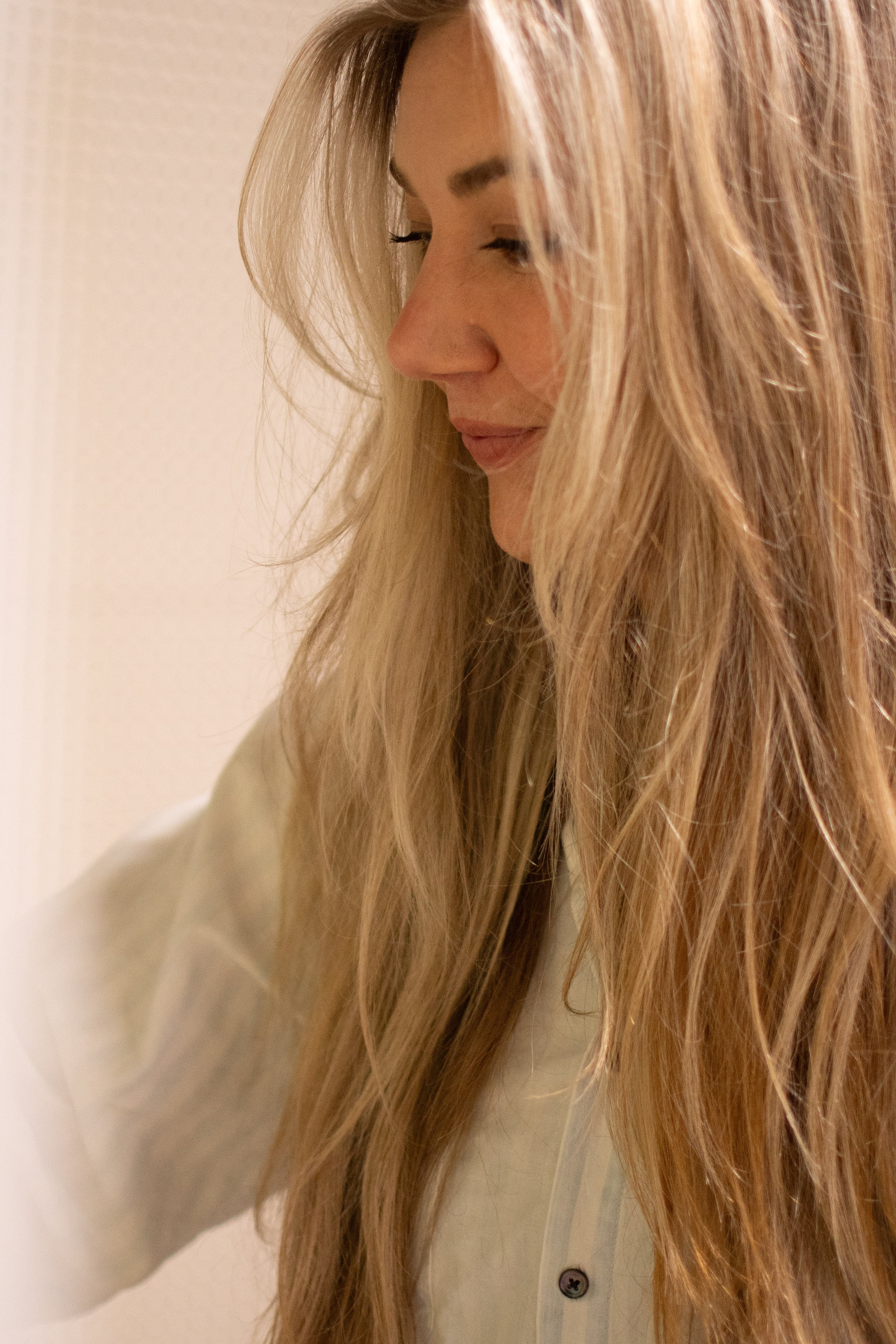 A close-up side profile of a woman with long, wavy blonde hair wearing a light-colored button-up shirt against a soft beige background.