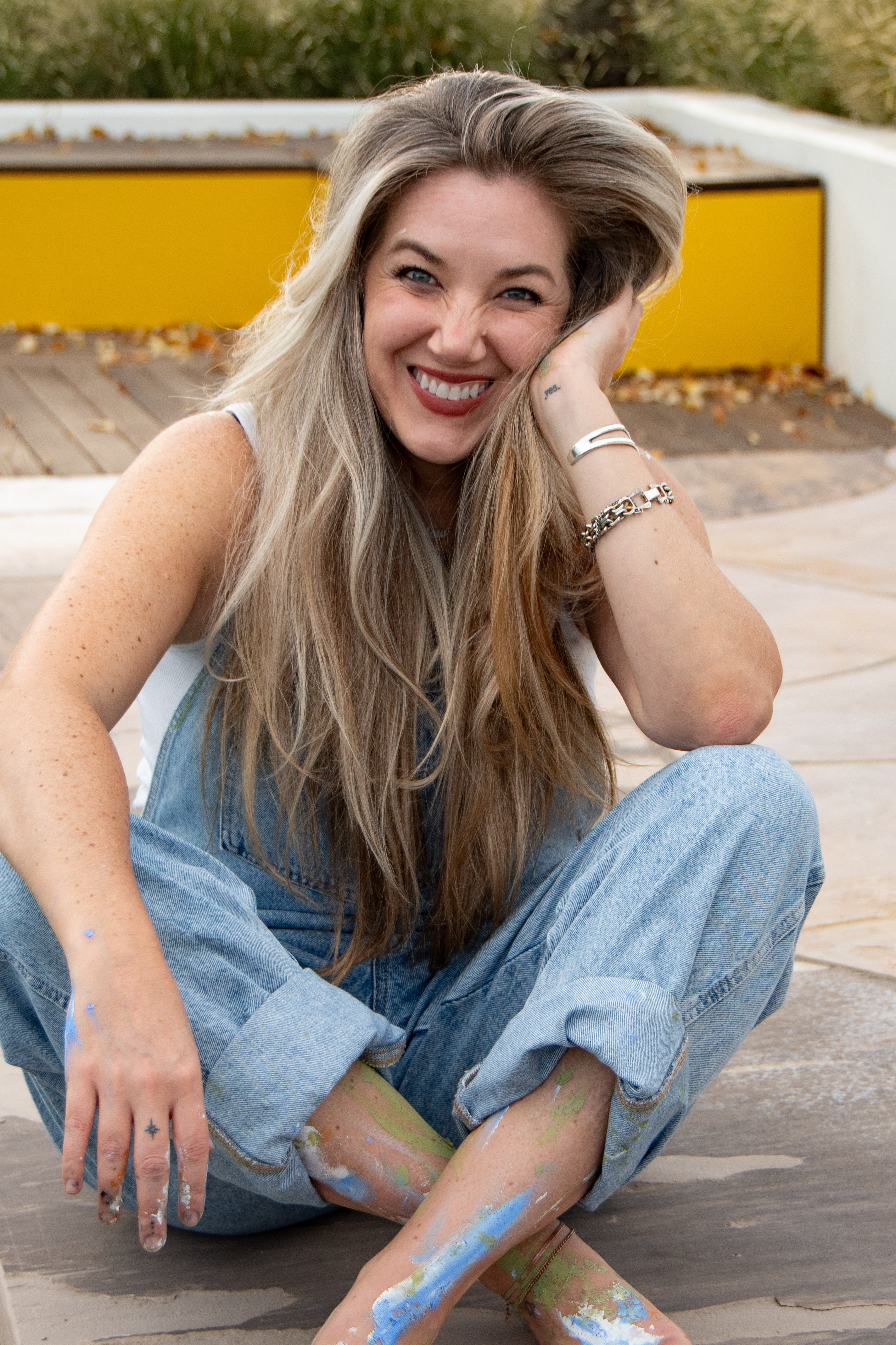 A smiling woman with long blonde hair sits outdoors on a wooden deck, wearing light blue denim overalls and a white sleeveless top. Paint is visible on her hands and legs, with a yellow planter and modern outdoor seating in the background.